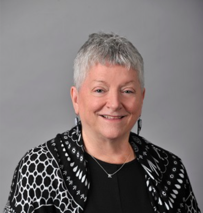 Headshot of a School of Social Work faculty member with short gray hair, smiling, wearing a black top and patterned cardigan against a neutral background.